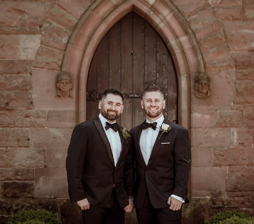 Two men in tuxedos standing in front of a rustic wooden door on a stone wall, smiling at the camera.