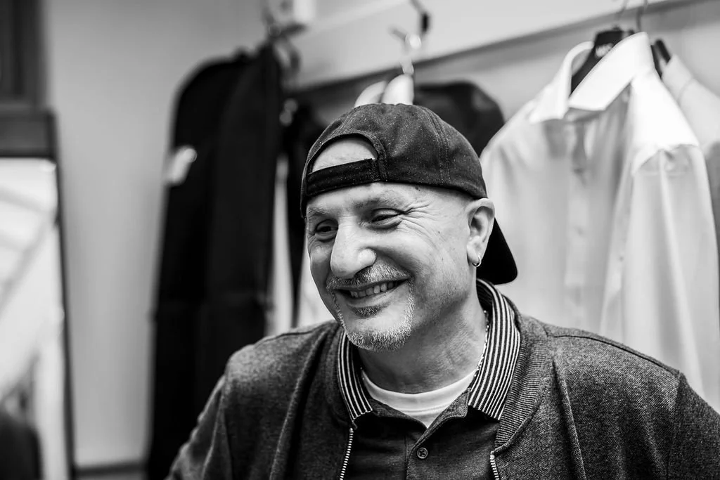 A smiling man wearing a backwards cap and a collared shirt, sitting in a room with hanging clothes in the background.