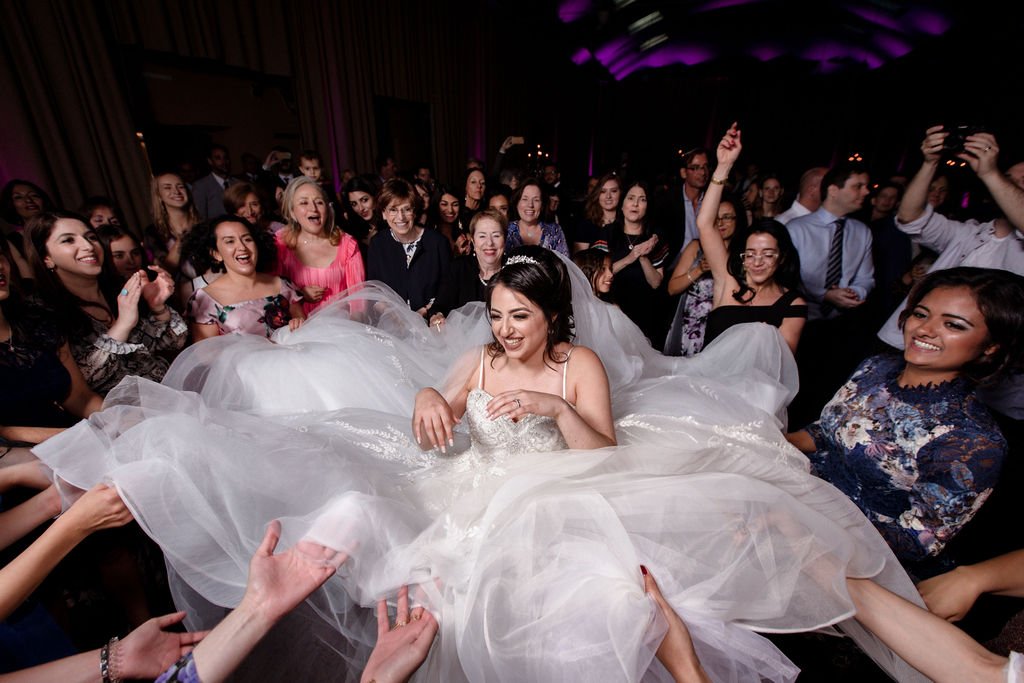 Bride in a white wedding gown being lifted by friends and family at her wedding reception, joyful and surrounded by smiling guests in a dimly lit venue with purple lighting.