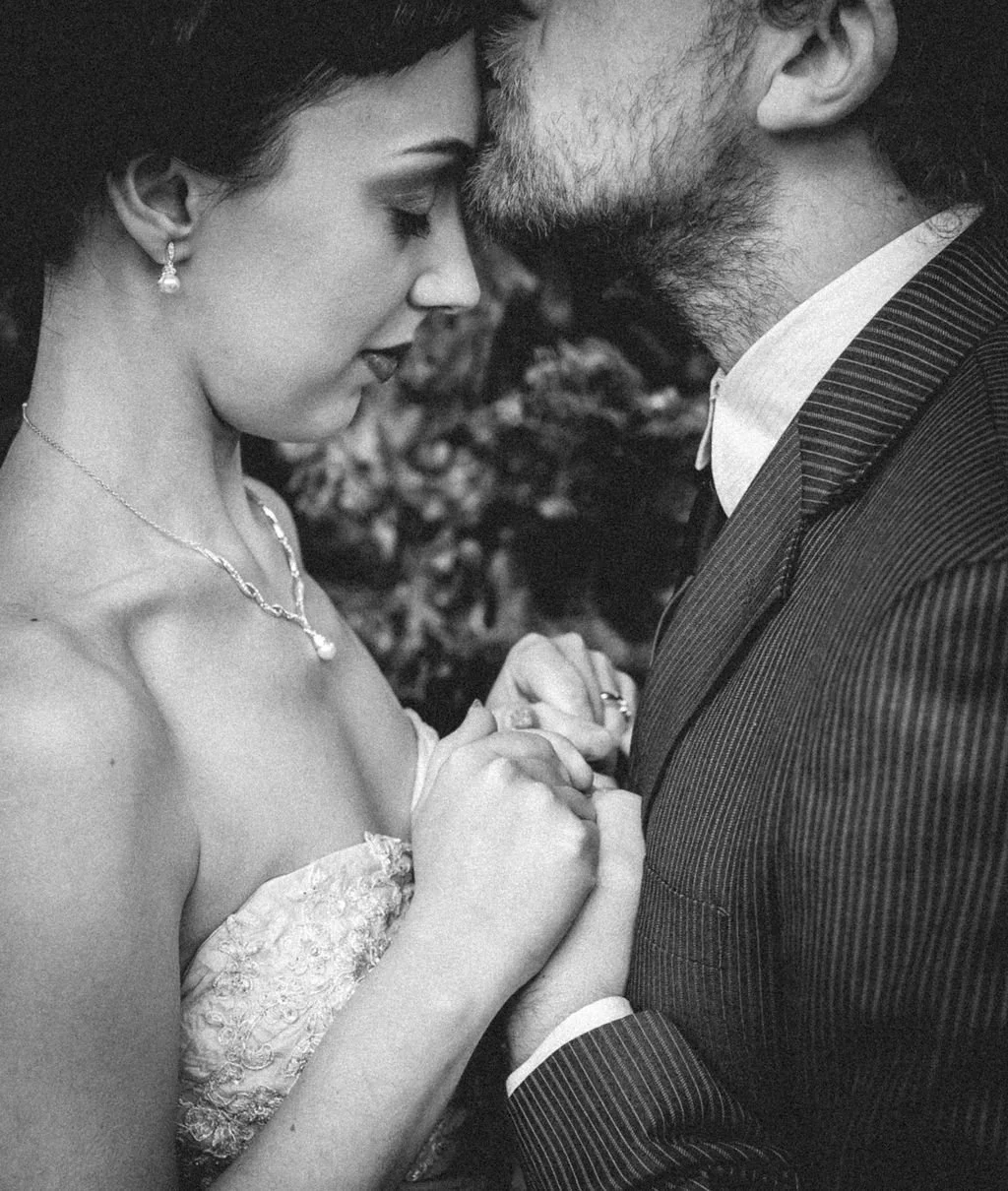 A black and white photo of a couple holding hands and touching foreheads, dressed formally, with the woman in an off-shoulder dress and jewelry, and the man in a pinstripe suit.