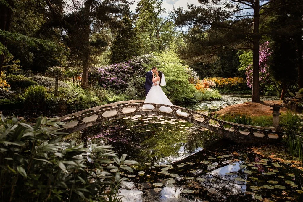 A bride and groom sharing a kiss on a small arched bridge over a reflective pond in a lush garden surrounded by blooming flowers and trees.