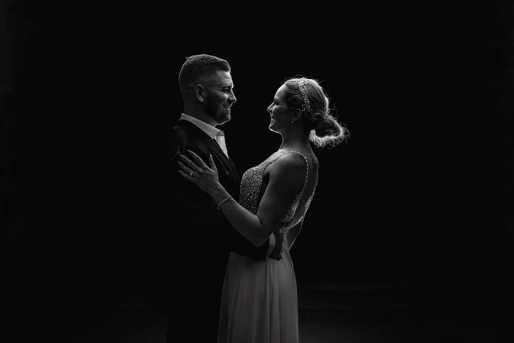 A black and white silhouette of a couple, a man and woman, embracing and smiling at each other against a dark background, possibly at a wedding or special event.