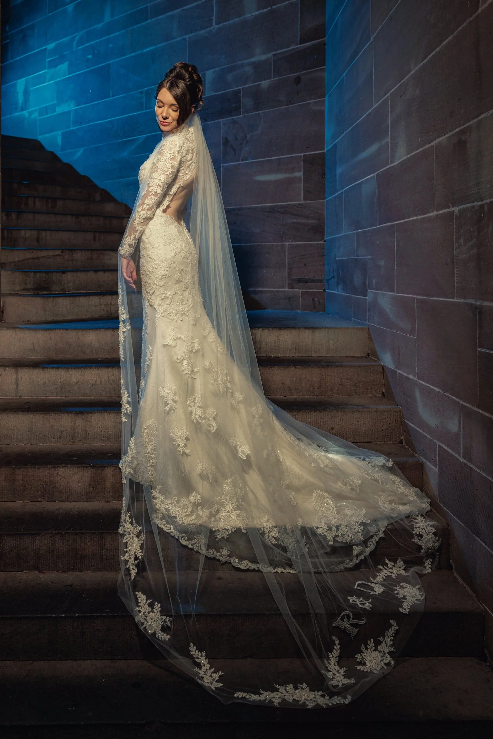 A bride in a lace wedding gown with a long train and sheer long sleeves, standing on a staircase against a dark stone wall, with her eyes closed and a serene expression.