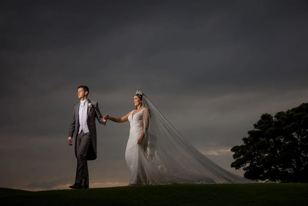 Bride and groom holding hands outdoors during cloudy sunset, with the bride in a white wedding dress and veil, and the groom in a gray tuxedo.