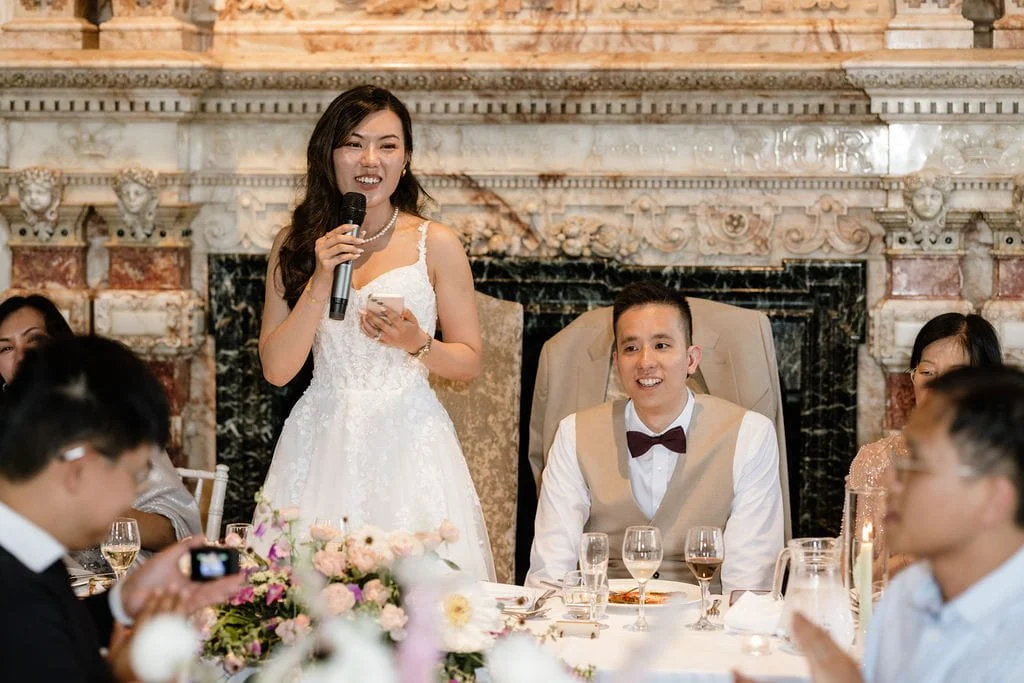 A woman in a white wedding dress is speaking into a microphone during a wedding reception, while a man in a beige vest and bow tie is seated at the table, listening and smiling. Other guests are sitting around the table with floral decorations and gl