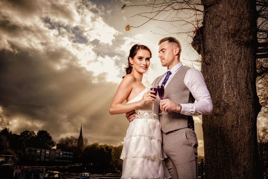 A bride and groom in wedding attire holding glasses of wine outdoors at sunset.