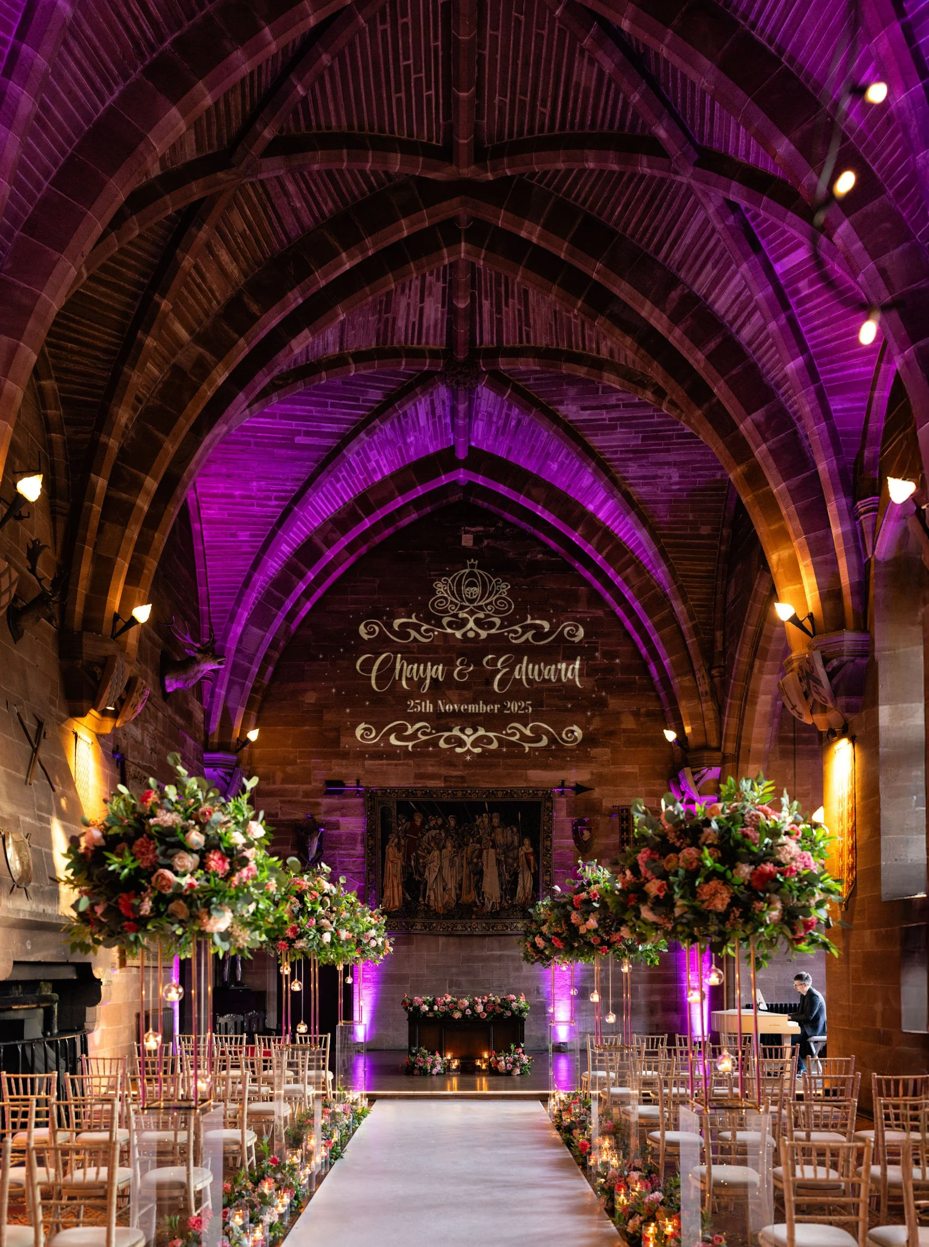 A decorated wedding venue in a church with floral arrangements and purple lighting. The altar has a sign with the names Chaya and Edward, and the date 25th November 2025.