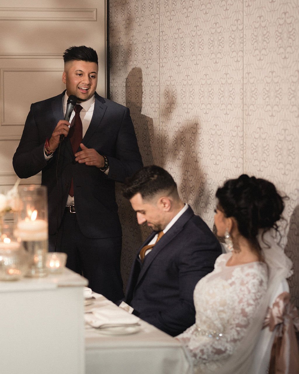 A man giving a speech with a microphone at a formal event, seated at a table with a woman in a lace dress and a man in a tuxedo.
