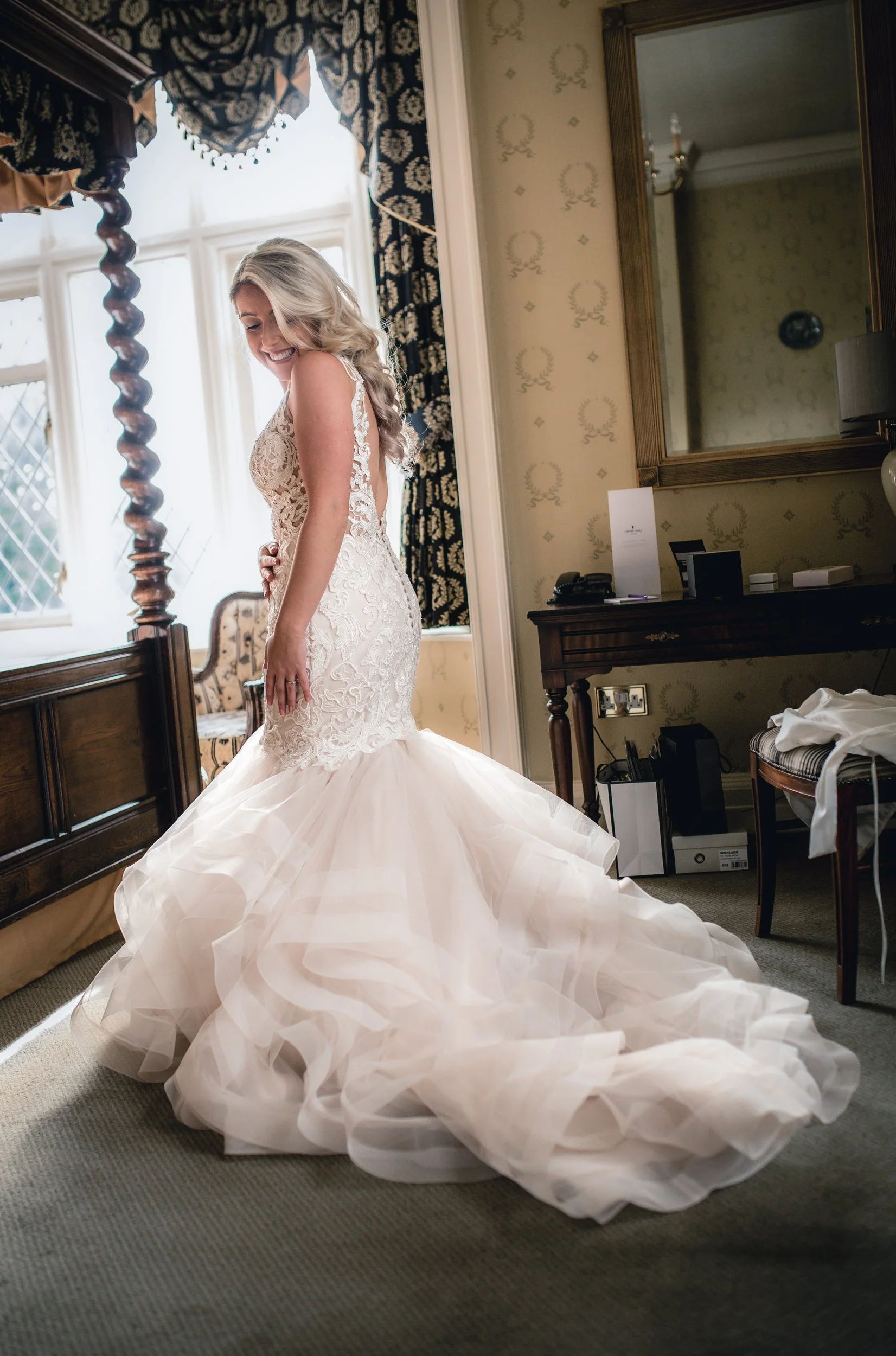 A bride in a lace and tulle wedding gown standing in a room with vintage decor, smiling and looking down.
