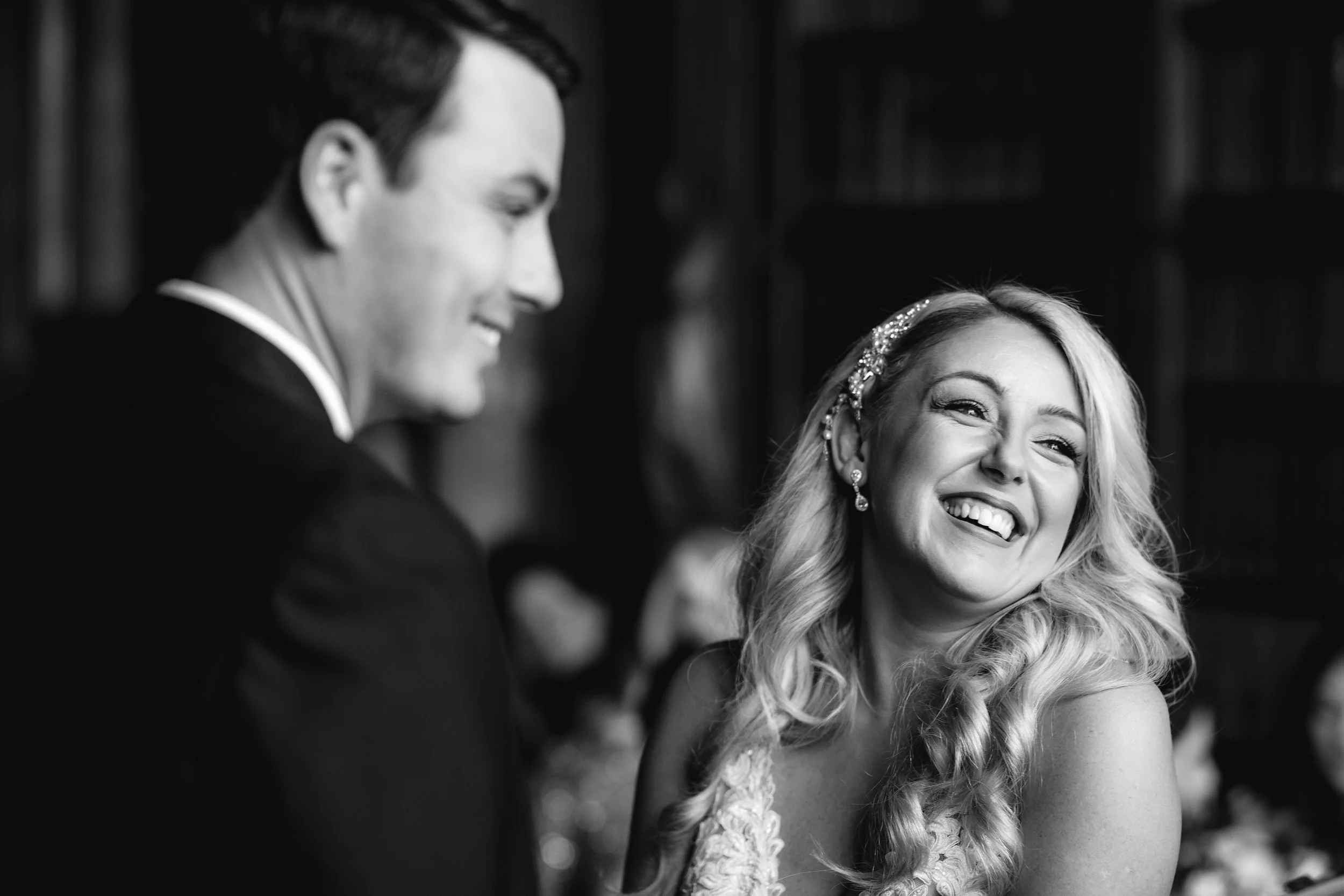 A black and white photo of a smiling woman with long, curly hair and wearing a decorative headband, looking at a man in a suit, at what appears to be a wedding or celebration.