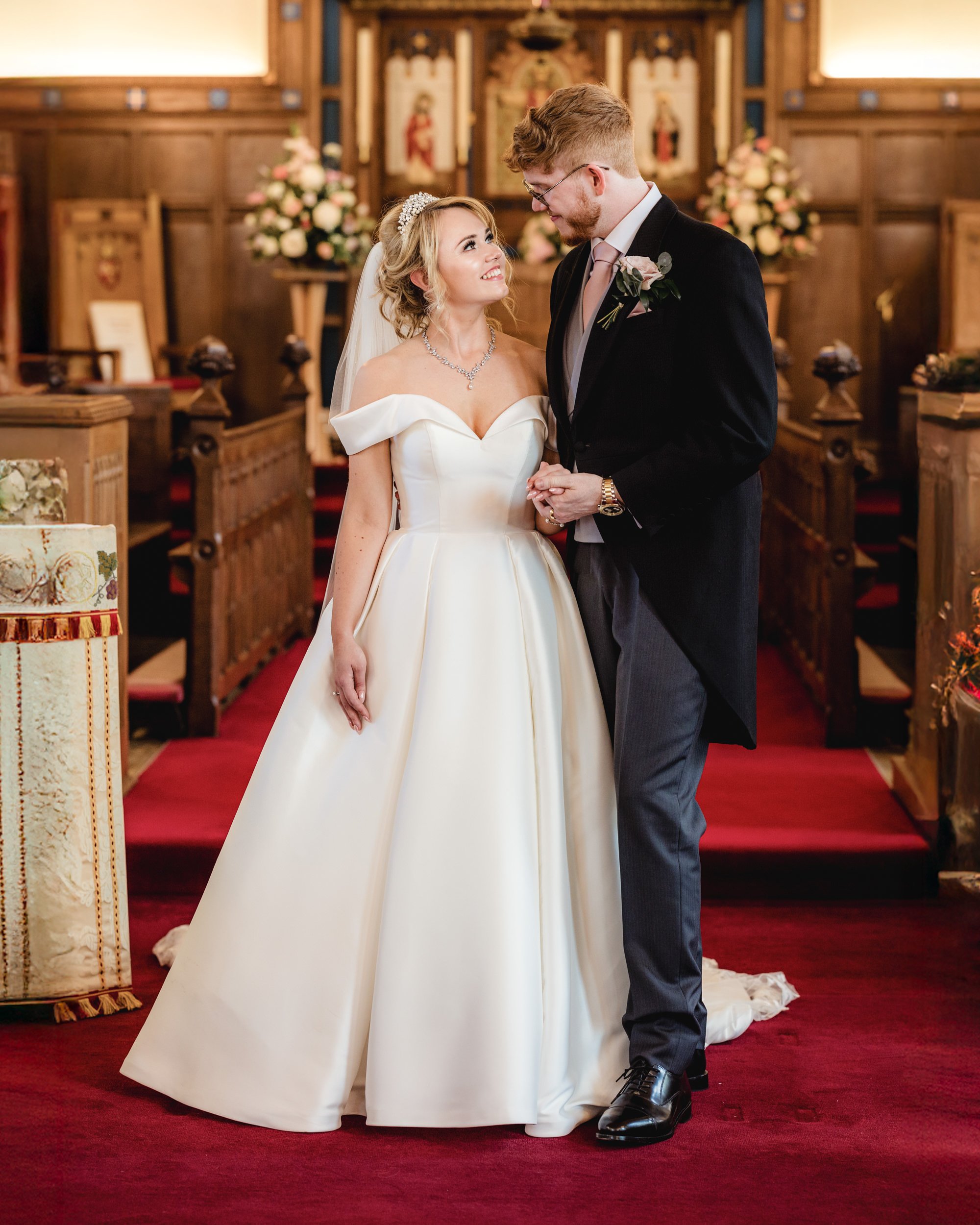 A bride and groom standing in a church during their wedding ceremony, holding hands and looking at each other happily. The bride is wearing a white off-shoulder wedding gown with a tiara and veil, and the groom is dressed in a black coat, gray pants,