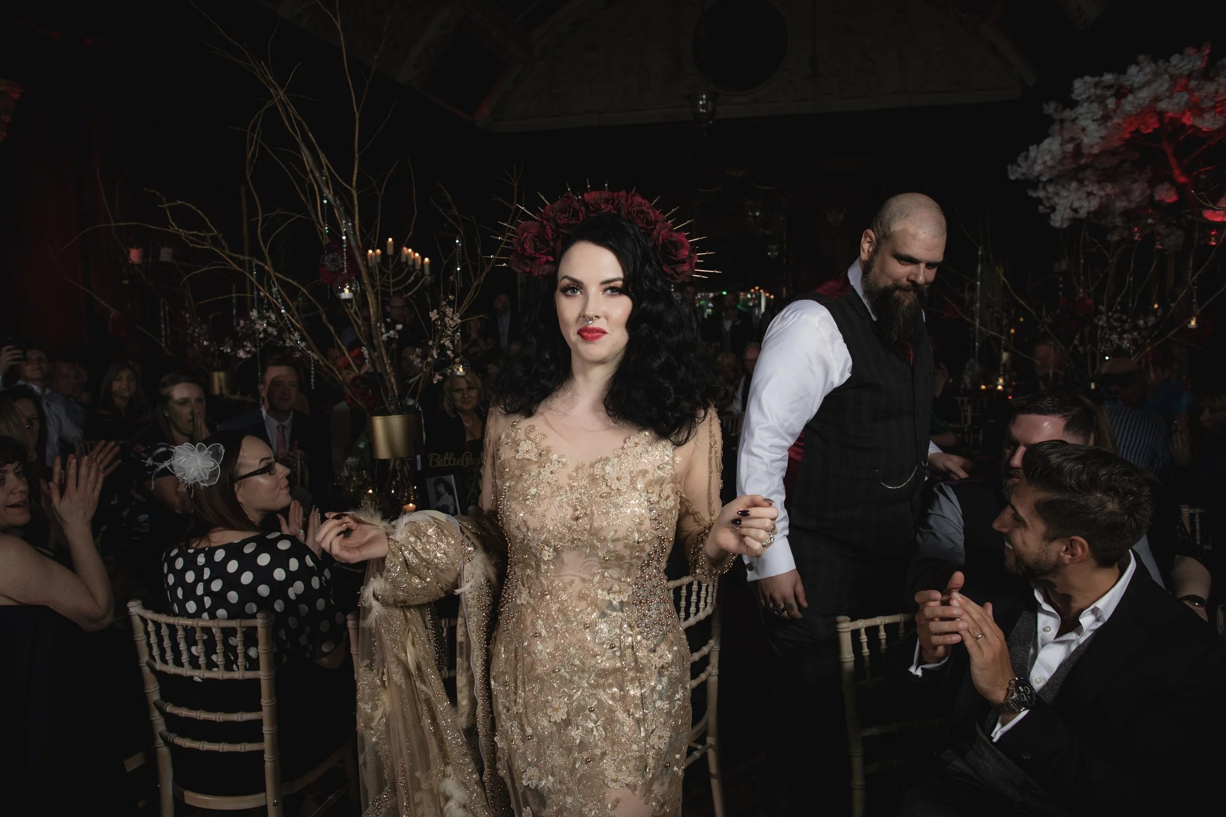 A woman with black wavy hair, wearing a gold embroidered gown and a headpiece made of dark red roses, walks through a decorated event space holding hands with a man with a beard in a vest. Several guests sit at tables, some clapping, in a dimly lit s