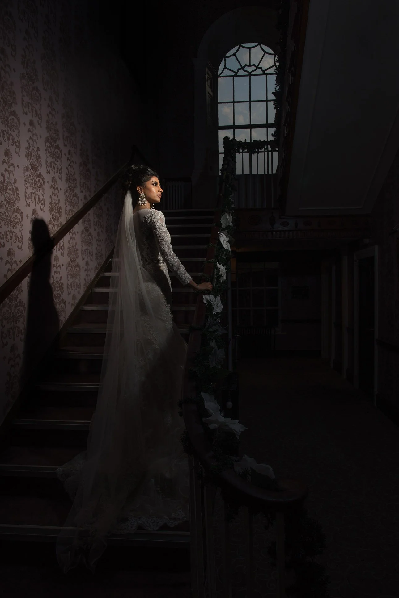 Beautifuk Asian bride in white dress stands on a staircase looking to one side in the evening with a window above her.