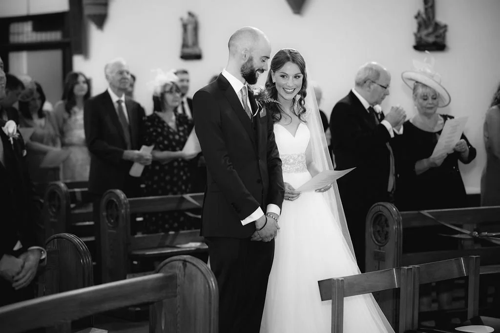 Black and white photo of a wedding ceremony showing a bride and groom standing together, with guests seated behind them in a church setting.