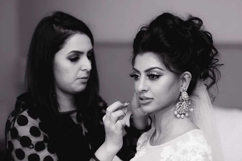 A woman with elaborate earrings is getting her makeup done by a makeup artist. The scene appears to be backstage or a preparation area for a special event, likely a wedding.
