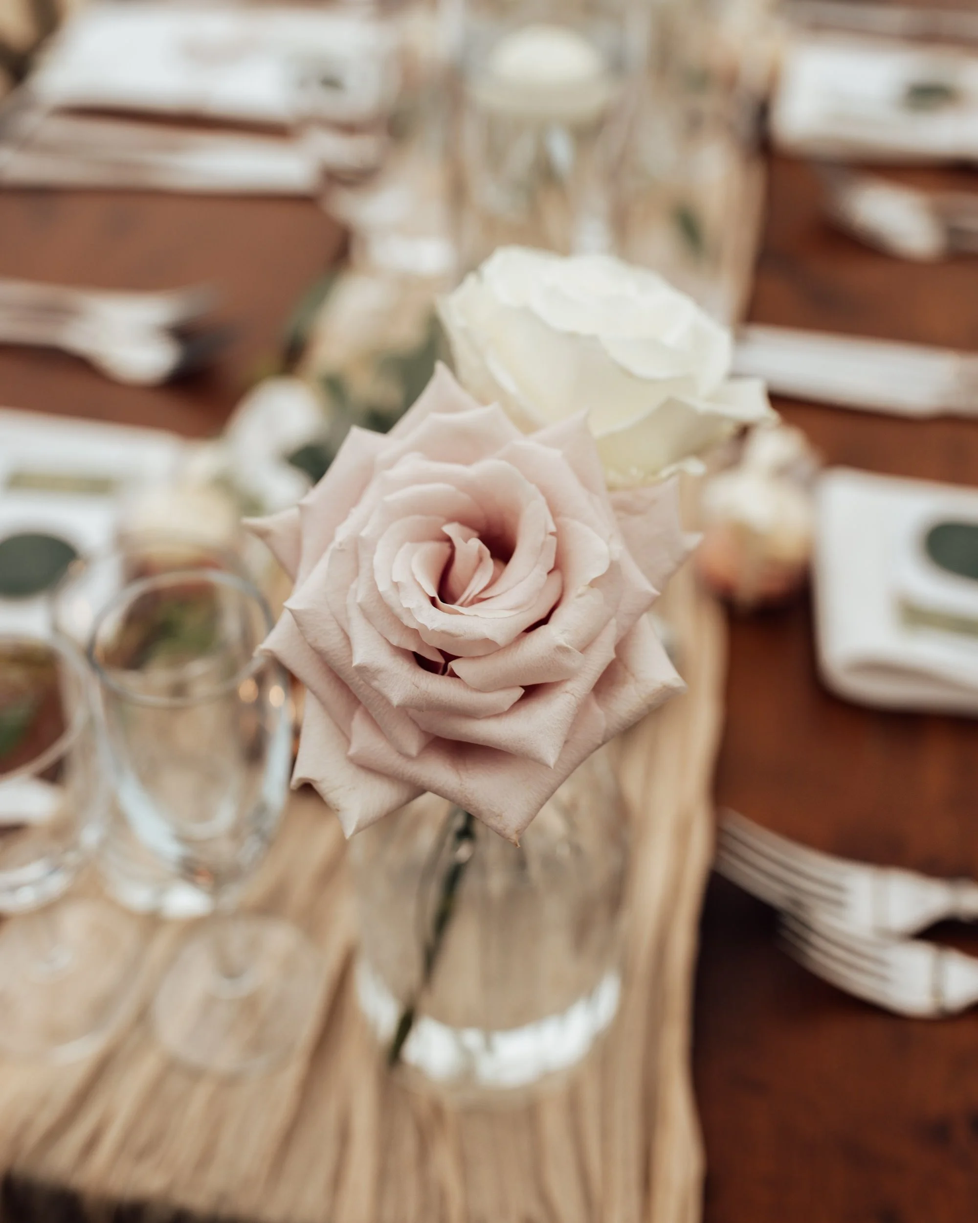 Close-up of pink and white roses on a rustic wooden table, with blank white plates and stacked napkins in the background, suggesting a wedding or formal event setup.