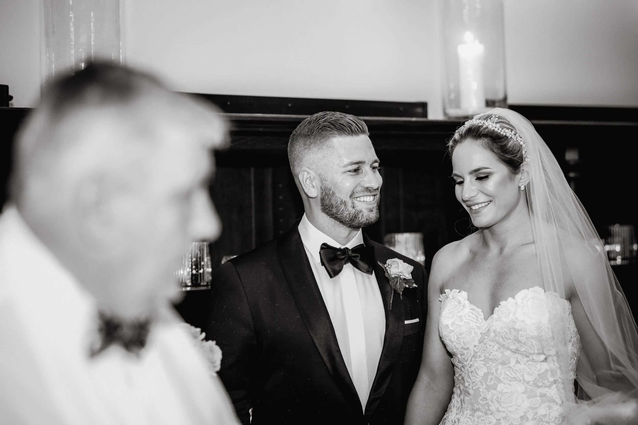 A black and white photo of a wedding ceremony with a bride and groom exchanging vows, smiling at each other, with a blurred officiant in the foreground.