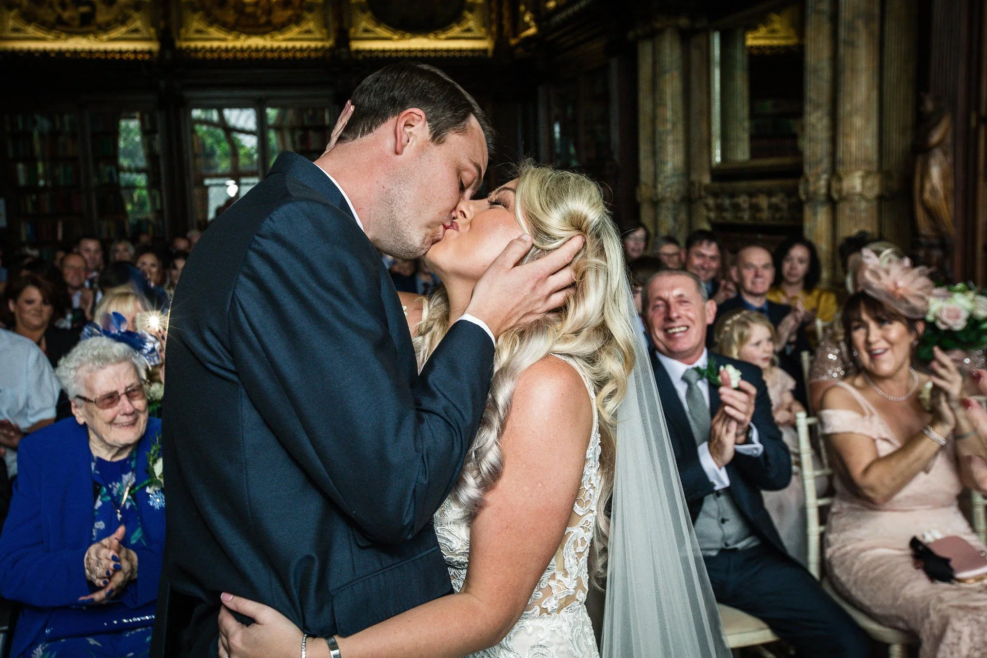 A couple sharing a kiss during their wedding ceremony, with guests clapping and smiling in the background.