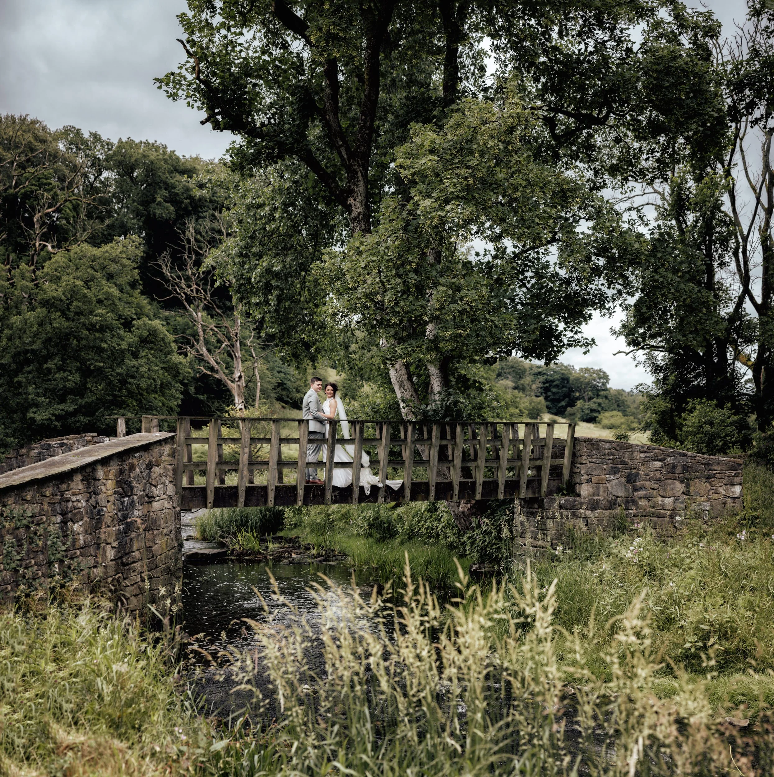 A bridge over a small stream in a wooded area, with a couple standing on the bridge, surrounded by lush green trees and grass.