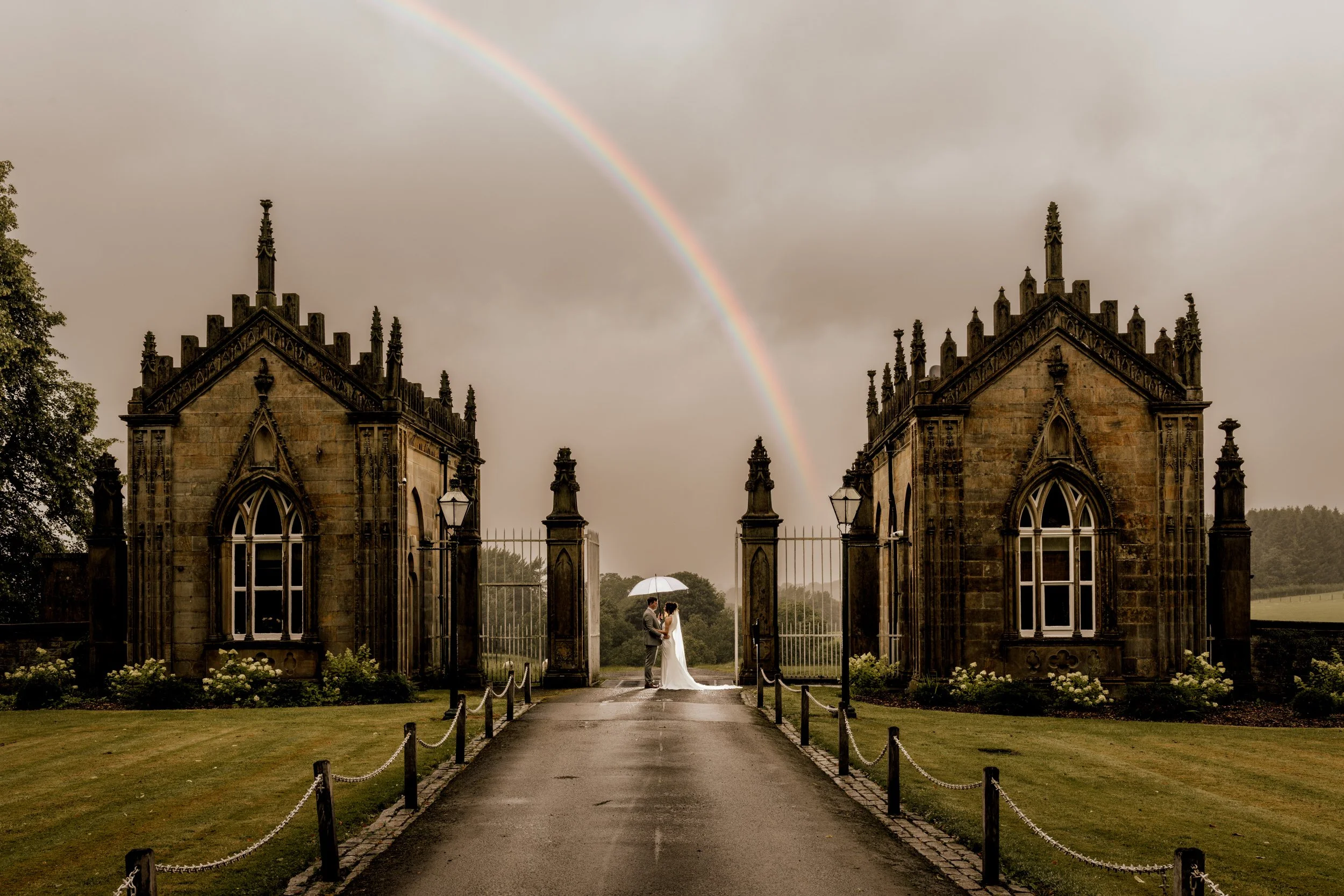 A couple in wedding attire stands under a rainbow near an ornate gothic-style gate with stone towers, during a rainy day.
