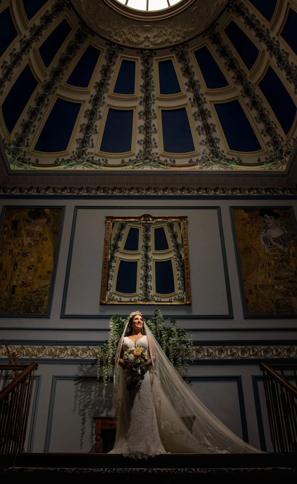 A bride in a white wedding gown and veil, holding a bouquet of flowers, standing on a staircase beneath a decorative ceiling with blue panels and intricate gold and floral details, in an ornate interior space.