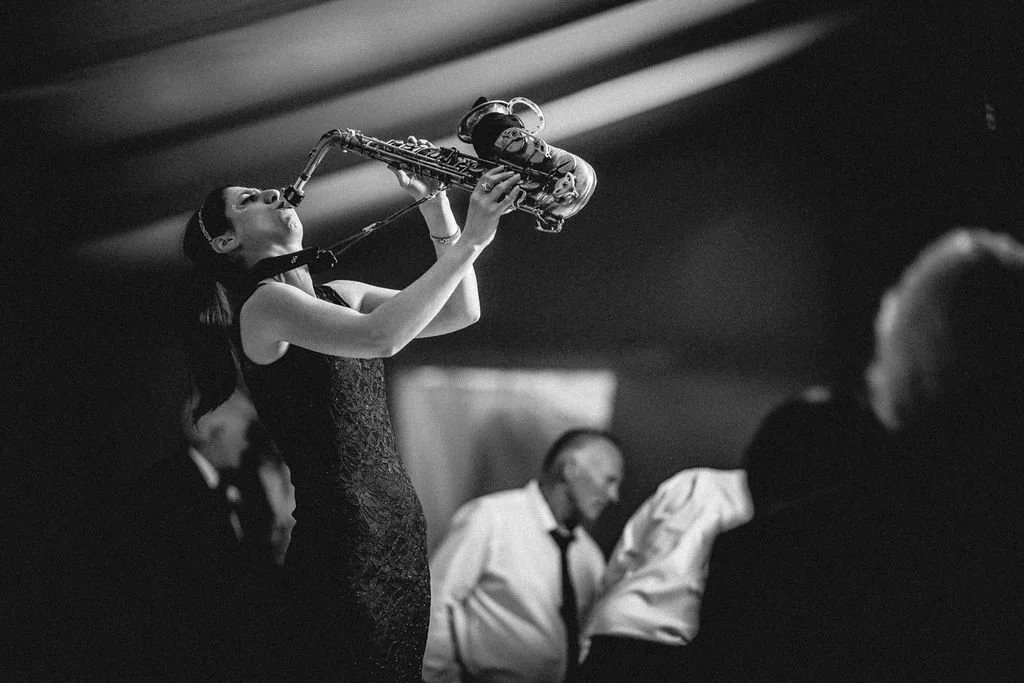 A woman in a sleeveless dress playing the saxophone on stage with other people in the background.