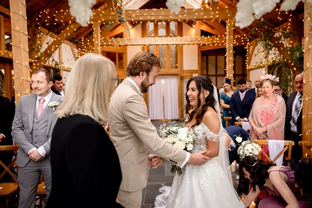 A bride and groom smiling and holding hands during their wedding ceremony, with guests seated behind them under a decorated wooden arch with string lights.
