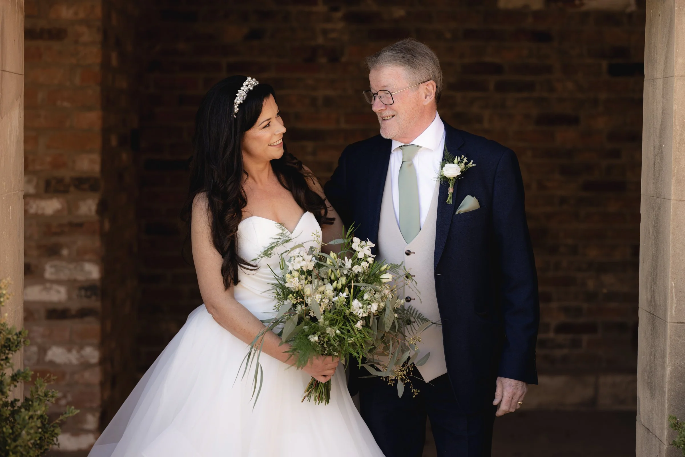 A bride and an older man, likely her father, sharing a happy moment during a wedding. The bride is holding a bouquet of white and green flowers, wearing a white wedding dress with a tulle skirt and a tiara. The man is dressed in a navy blue suit with