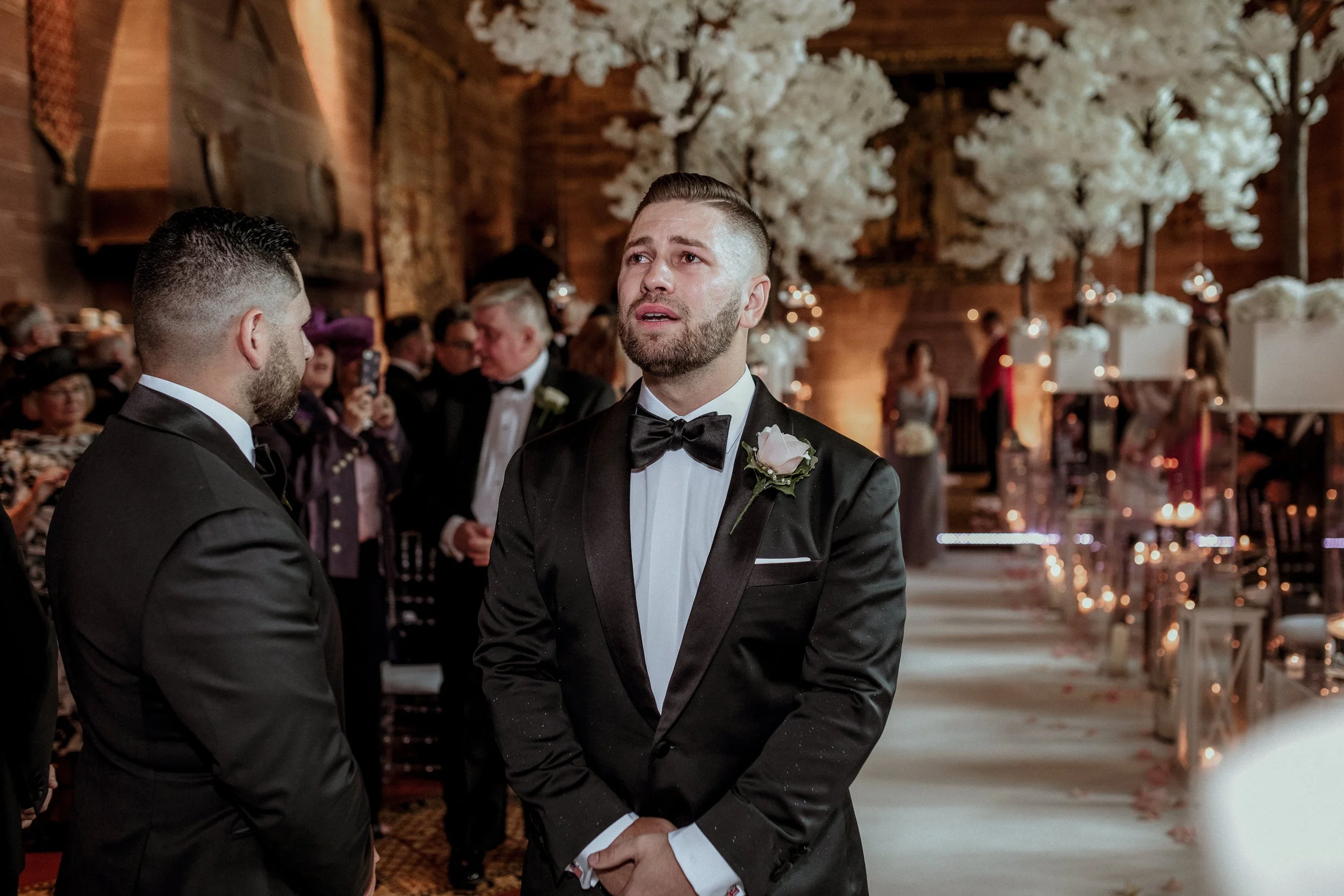 Groom with a beard and slicked-back hair wearing a black tuxedo and bow tie, standing at a wedding ceremony inside a decorated venue with white flowers and guests in formal attire.