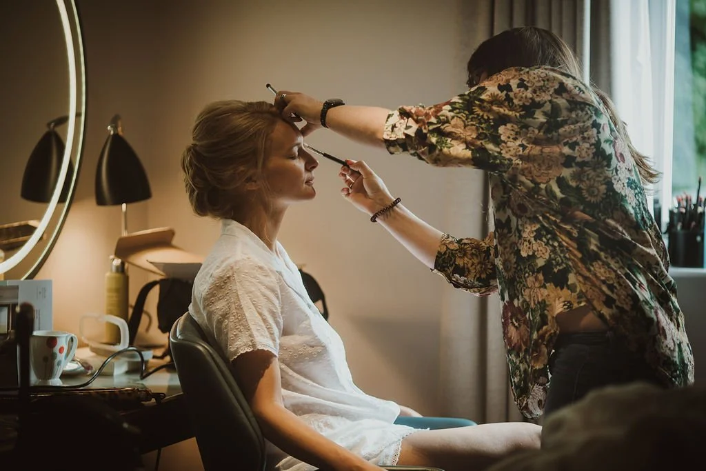 A woman getting her makeup or eyebrows done by a makeup artist in a bedroom.