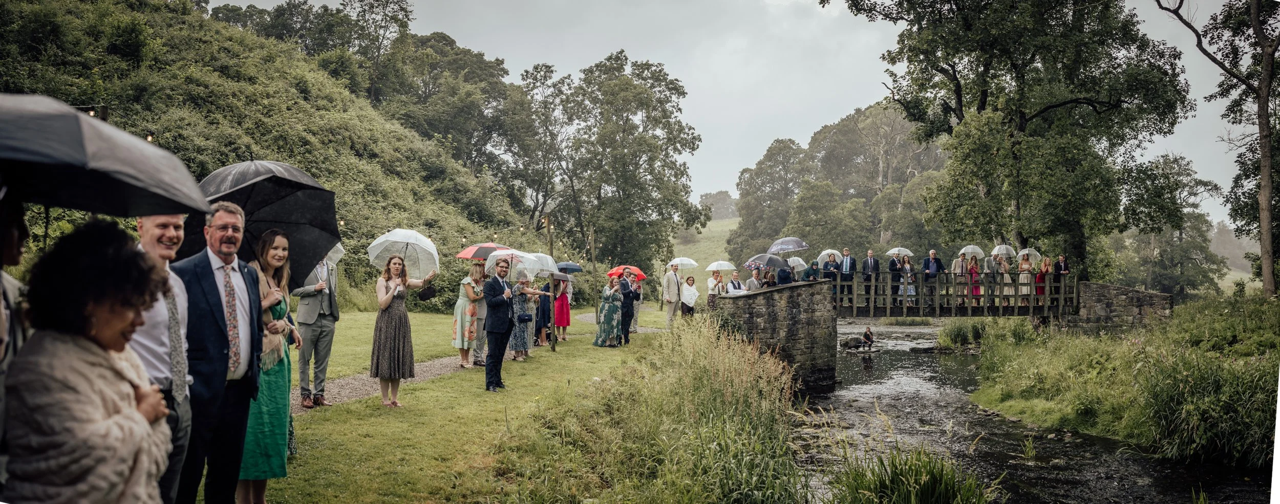 Group of wedding guests holding umbrellas outdoors near a small stone bridge over a creek on a rainy day, surrounded by trees and greenery.