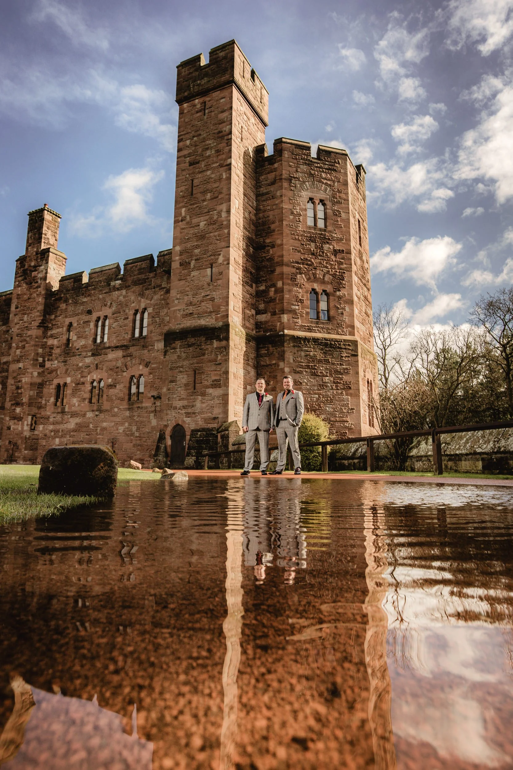 Two men in suits standing outside a castle with a water reflection in the foreground.