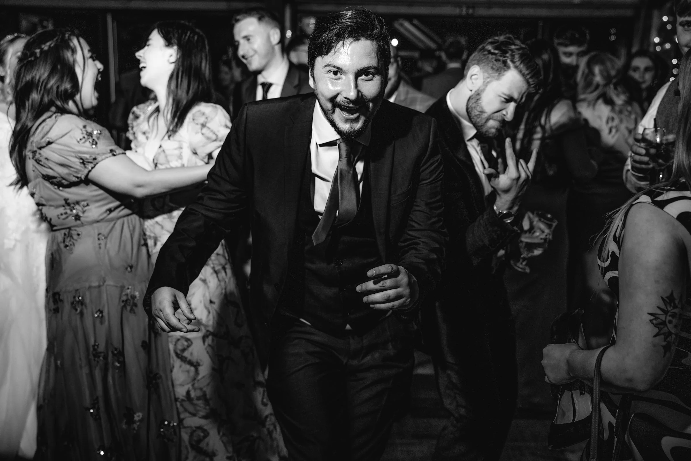 Black and white photo of a man in a tuxedo dancing and smiling at a lively party with other people dancing and socializing.