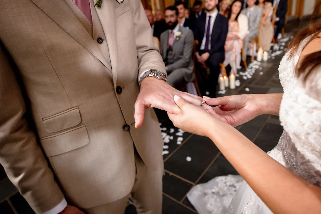 A bride and groom exchanging wedding rings during their ceremony, with guests seated in the background and candles on the aisle.