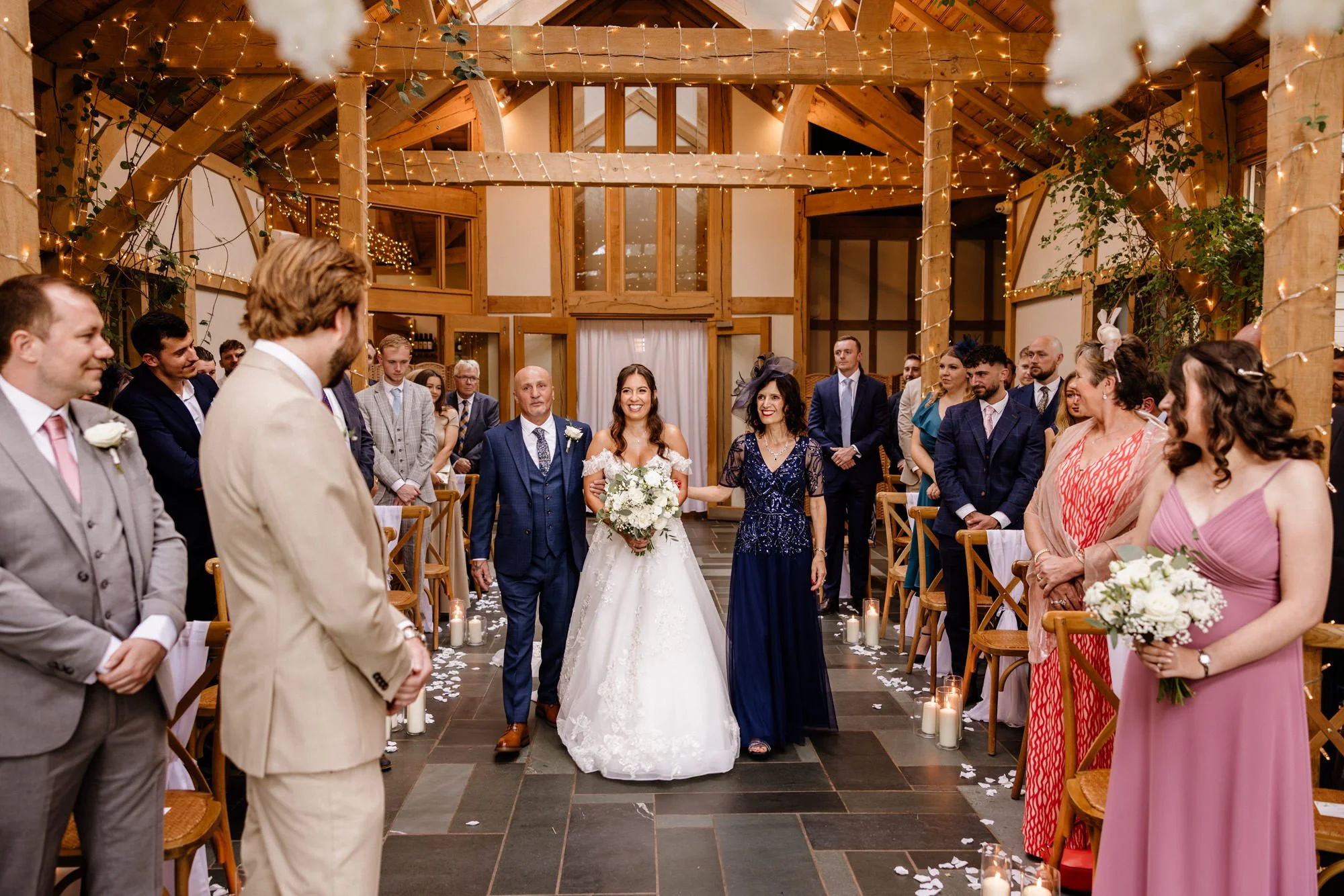 A wedding ceremony taking place indoors with guests standing and watching the bride walk down the aisle, decorated with candles and string lights, with a wooden interior and guests dressed in formal attire.