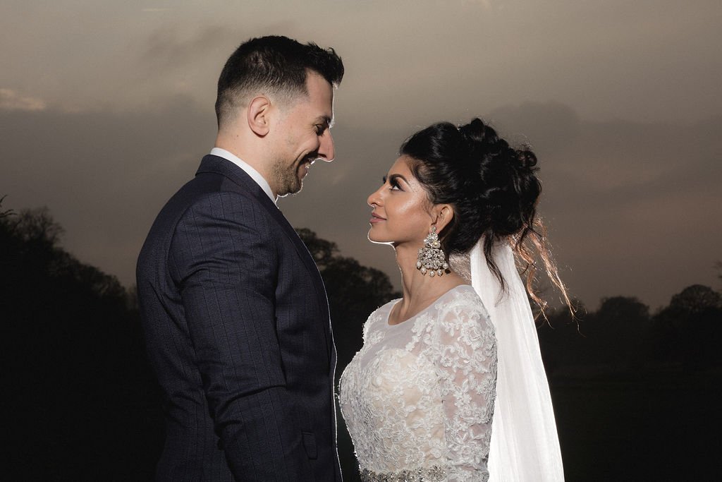 A bride and groom standing close together outdoors at sunset, smiling at each other with a dark sky and trees in the background.