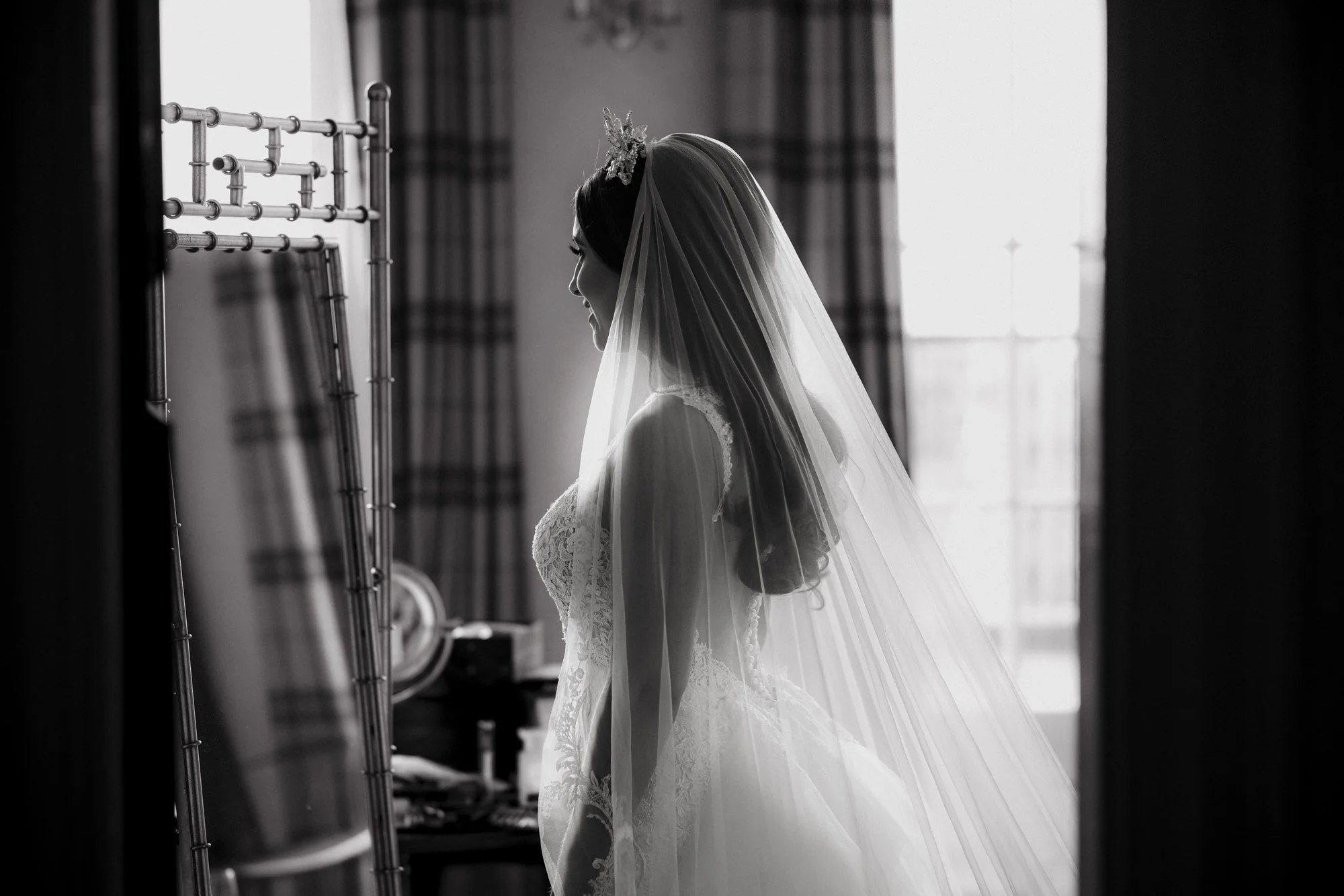 A bride in a wedding gown and veil standing indoors, looking at herself in a mirror, with natural light coming through a window in the background.