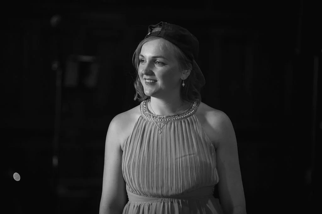 Black and white photo of a smiling woman wearing a dress with a beaded neckline, earrings, and a cap, standing indoors.