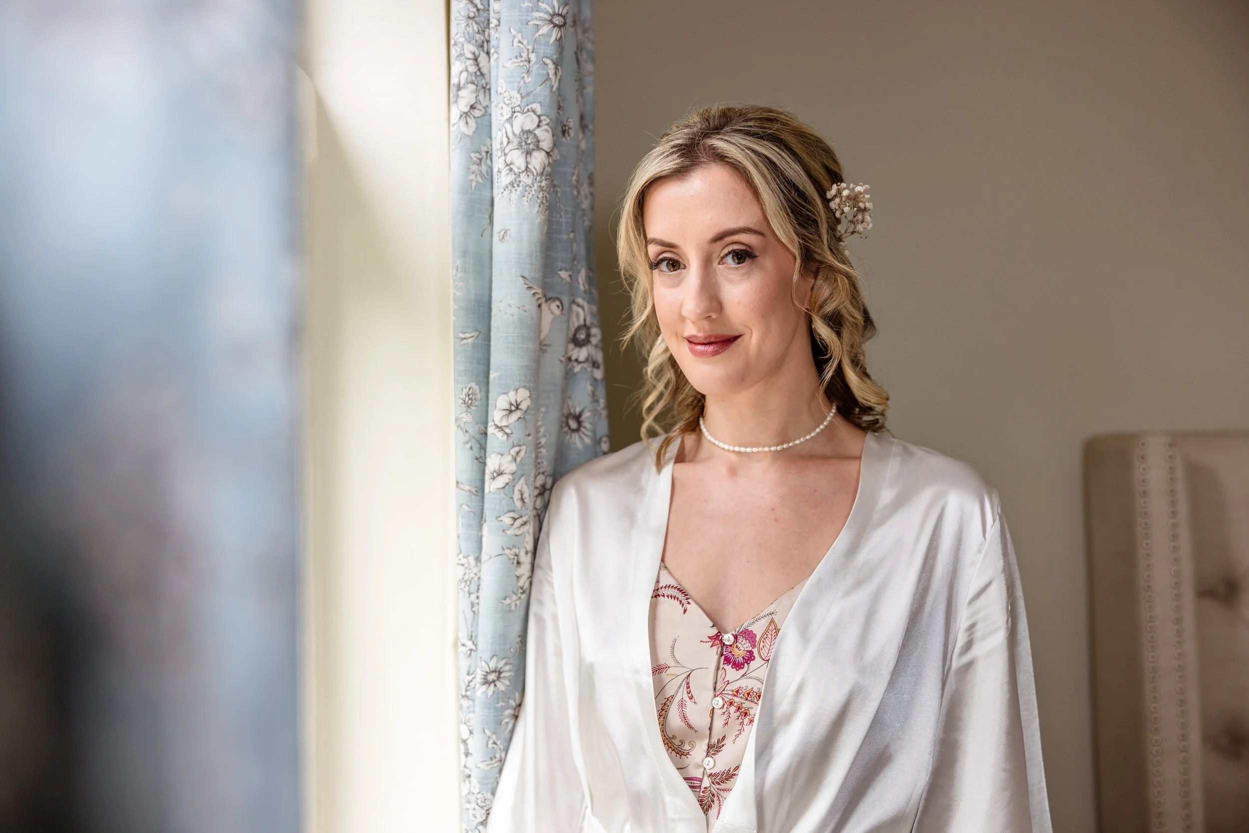 A woman dressed in a light satin robe and floral top, wearing a pearl necklace, standing near a window with floral curtains, smiling softly at the camera.