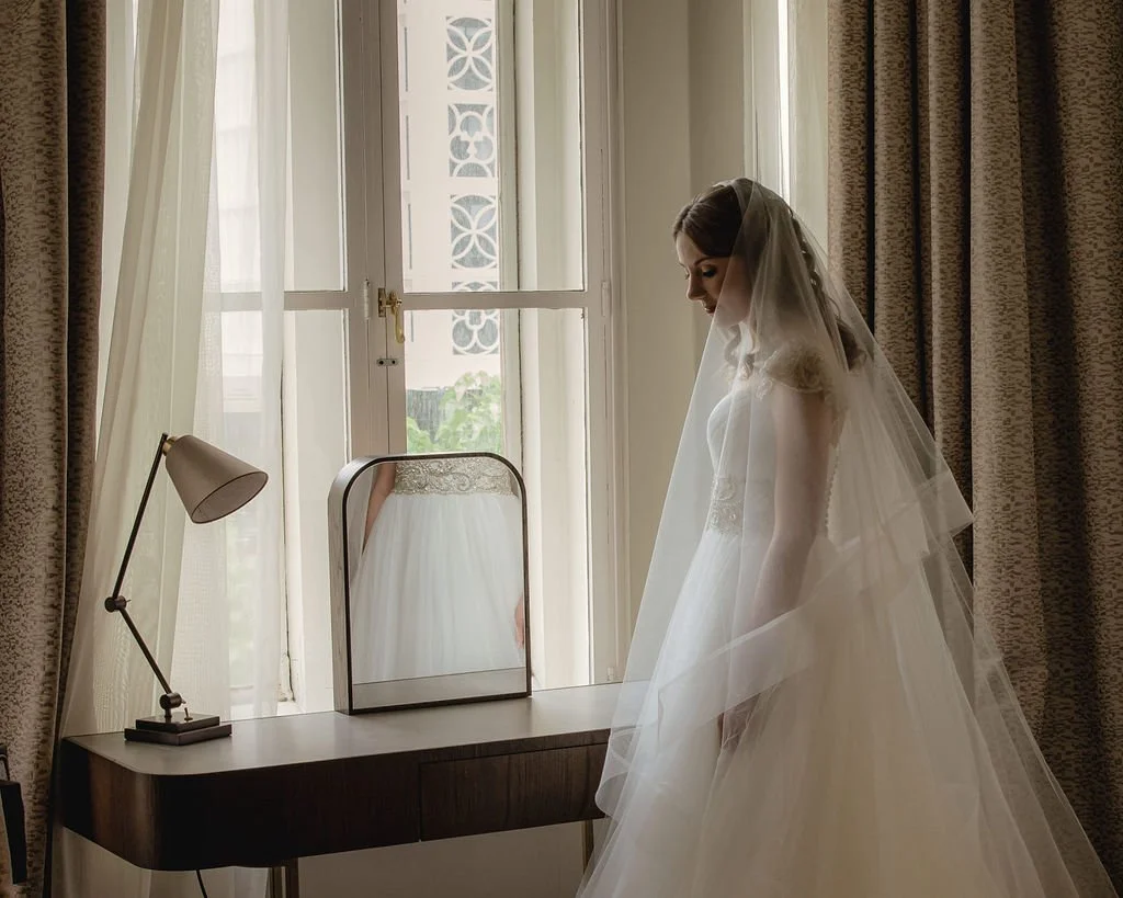 A bride in a wedding dress and veil standing by a window, looking down, with her reflection visible in a small mirror on a dresser.