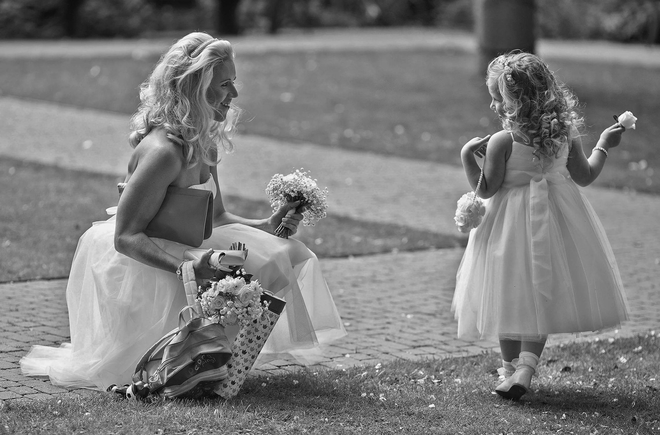 A woman in a wedding dress kneeling on the grass and smiling at a young girl in a dress with a bow on her back, as they exchange flowers in a park.