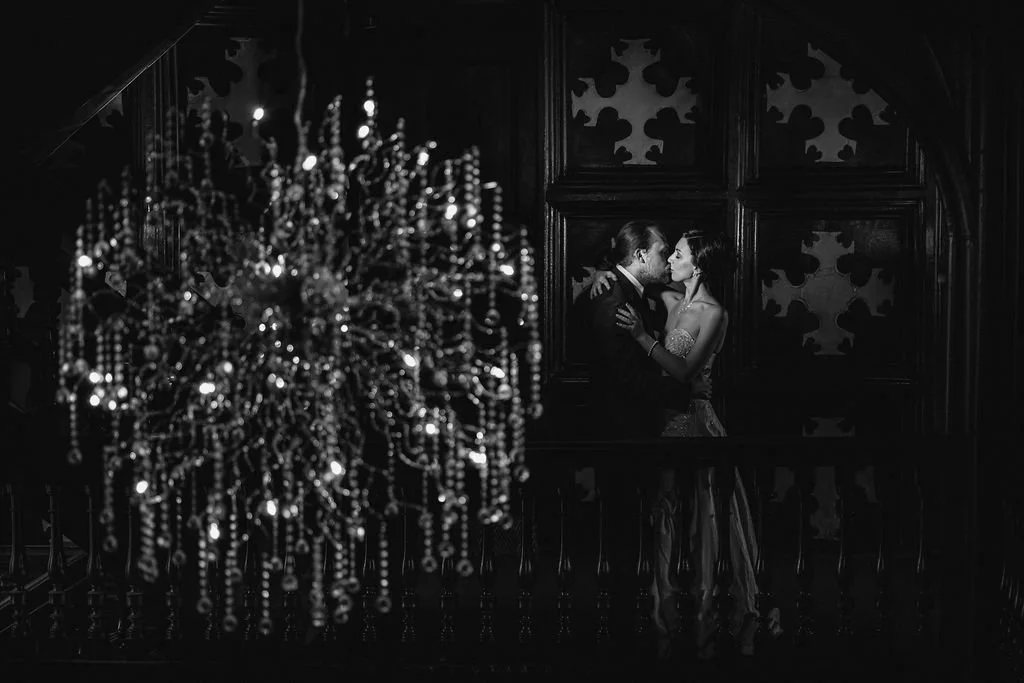 A black-and-white photo of a romantic couple dancing closely, with a large chandelier in the foreground, in a dimly lit elegant room with patterned walls.