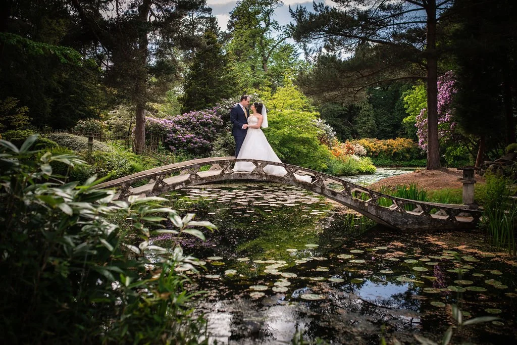 A bride and groom stand on a small arched stone bridge over a pond in a lush garden, surrounded by trees and colorful flowers.