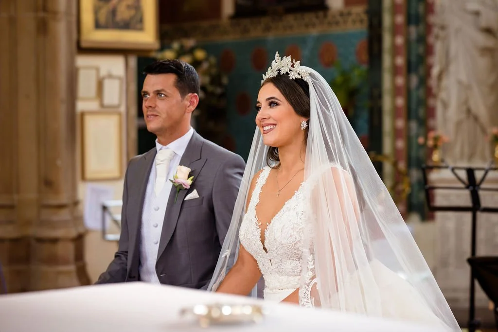 A bride and groom standing side by side during their wedding ceremony inside a church. The bride is smiling, wearing a white wedding dress with lace details, a veil, and a floral crown. The groom is in a gray suit with a white tie and pink boutonnier