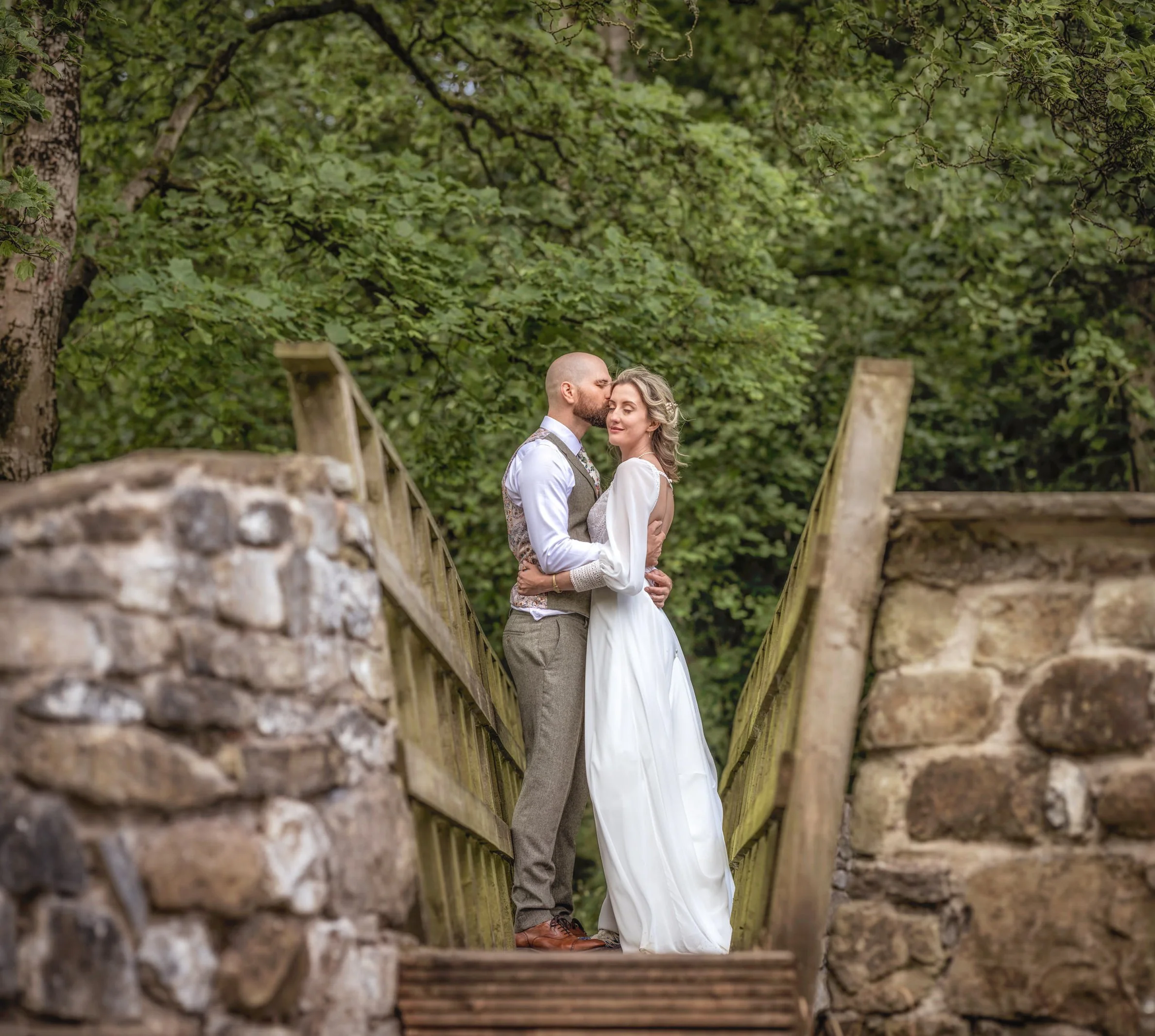 A couple in wedding attire embracing on a small wooden bridge in a lush, green forest.