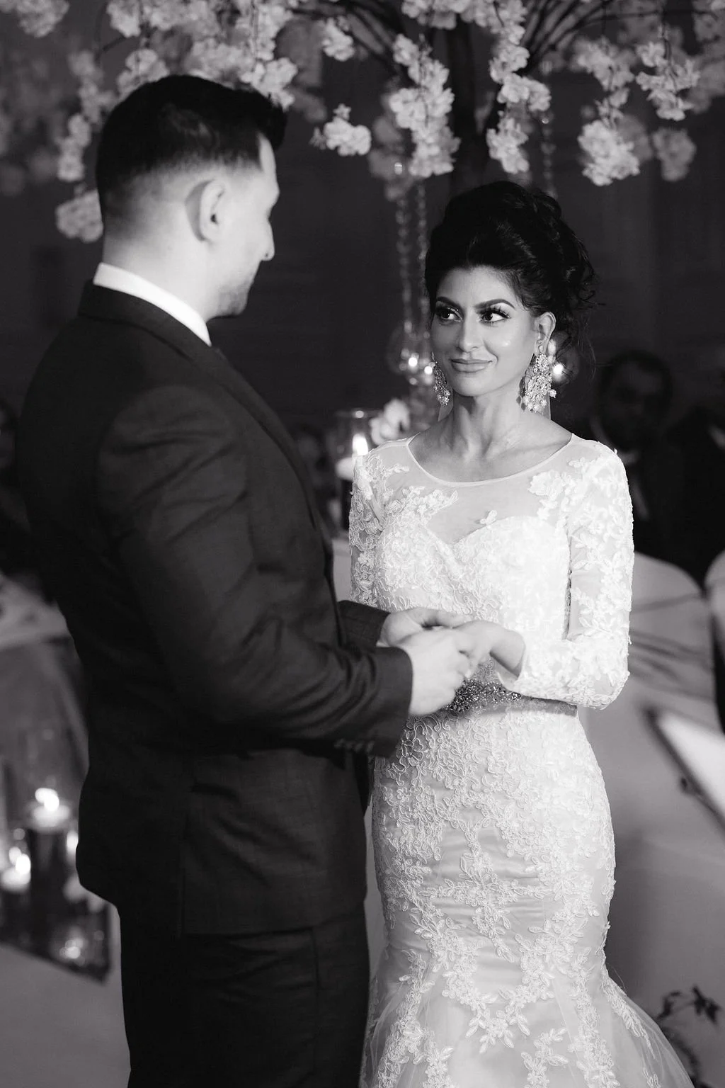 A couple exchanging vows during a wedding ceremony. The bride wears a lace wedding gown and large earrings, and the groom is dressed in a dark suit. They are holding hands and looking at each other, with floral decorations in the background.
