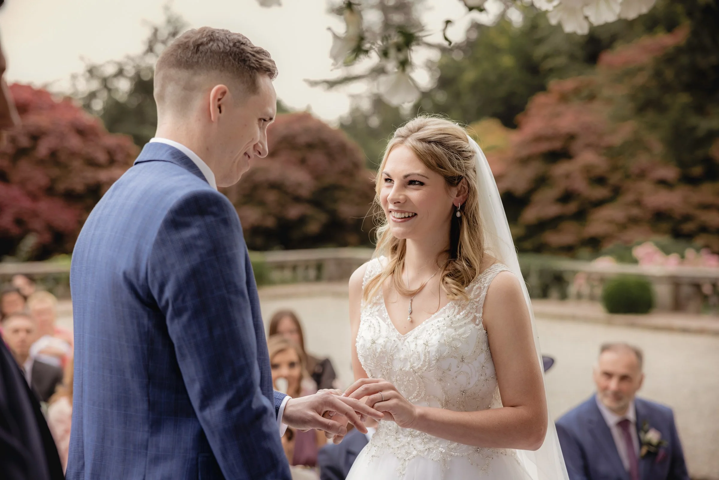 A bride and groom exchange wedding rings outdoors surrounded by guests and colorful trees.