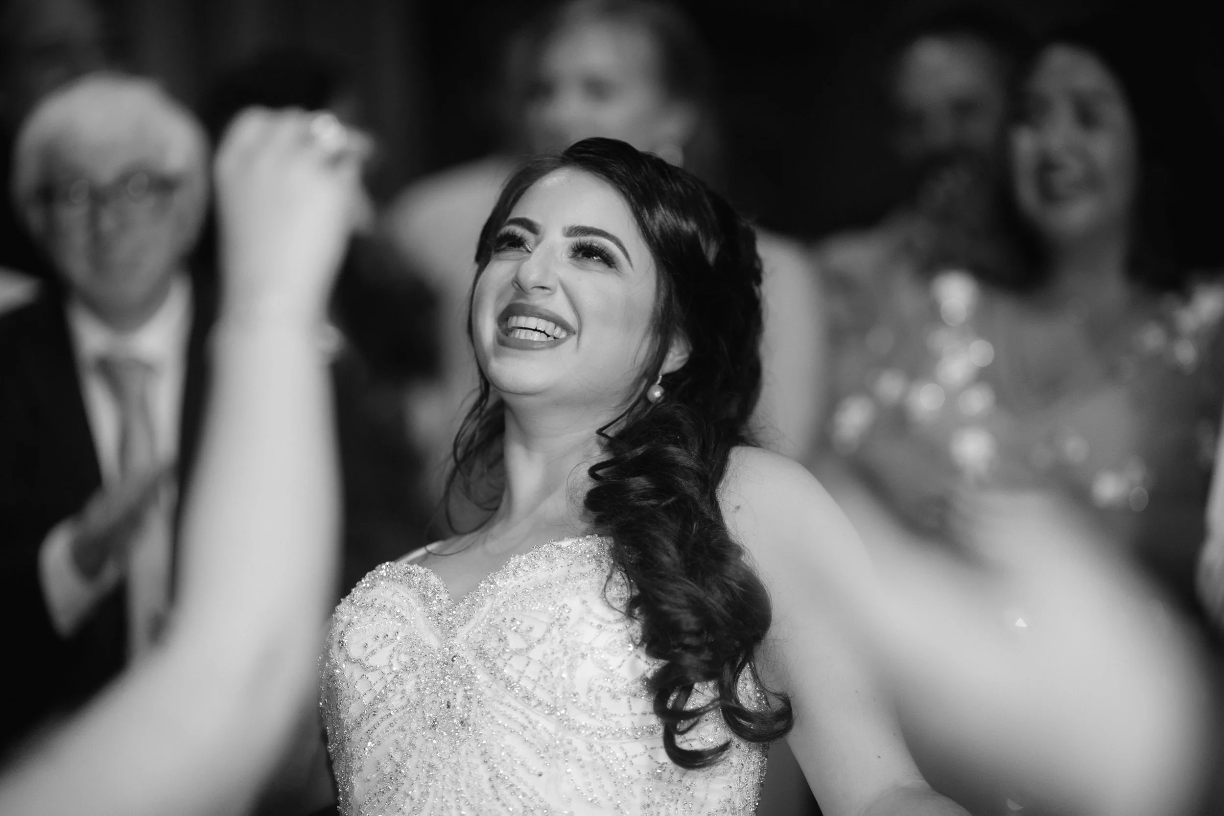 Black and white photo of a woman in a wedding dress smiling and looking up, with blurred people in the background at a celebration.