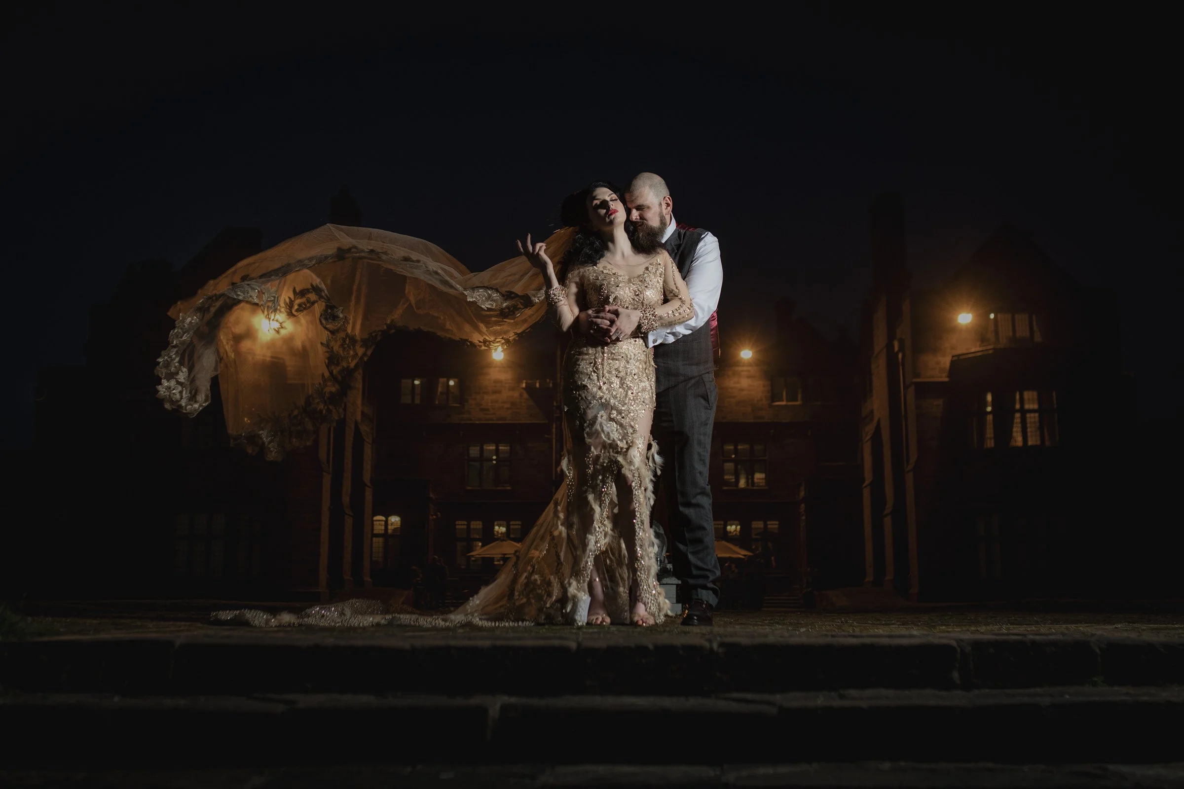 A couple dressed in formal attire standing close together outside at night, with a large historic building and glowing lights in the background.