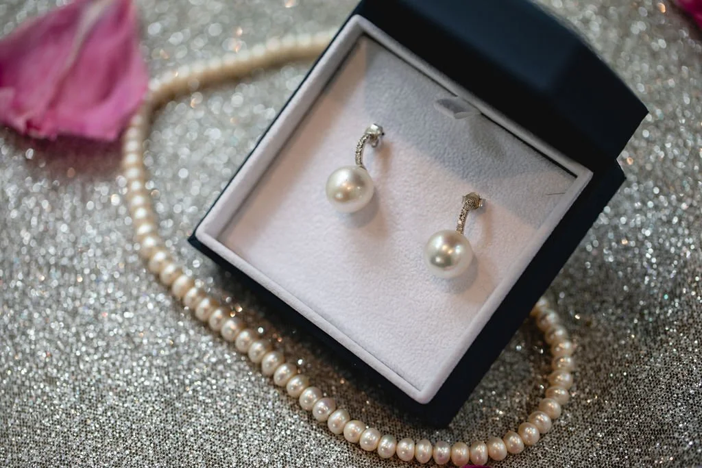 Pearl earrings inside a black jewelry box, with a pearl necklace and a pink flower petal on a glittery surface.